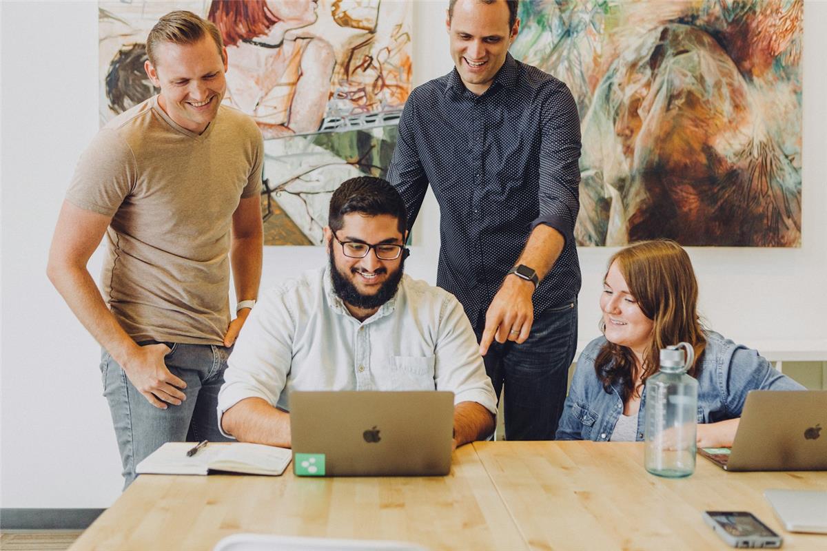 Four colleagues gathered around a laptop in a bright office, smiling